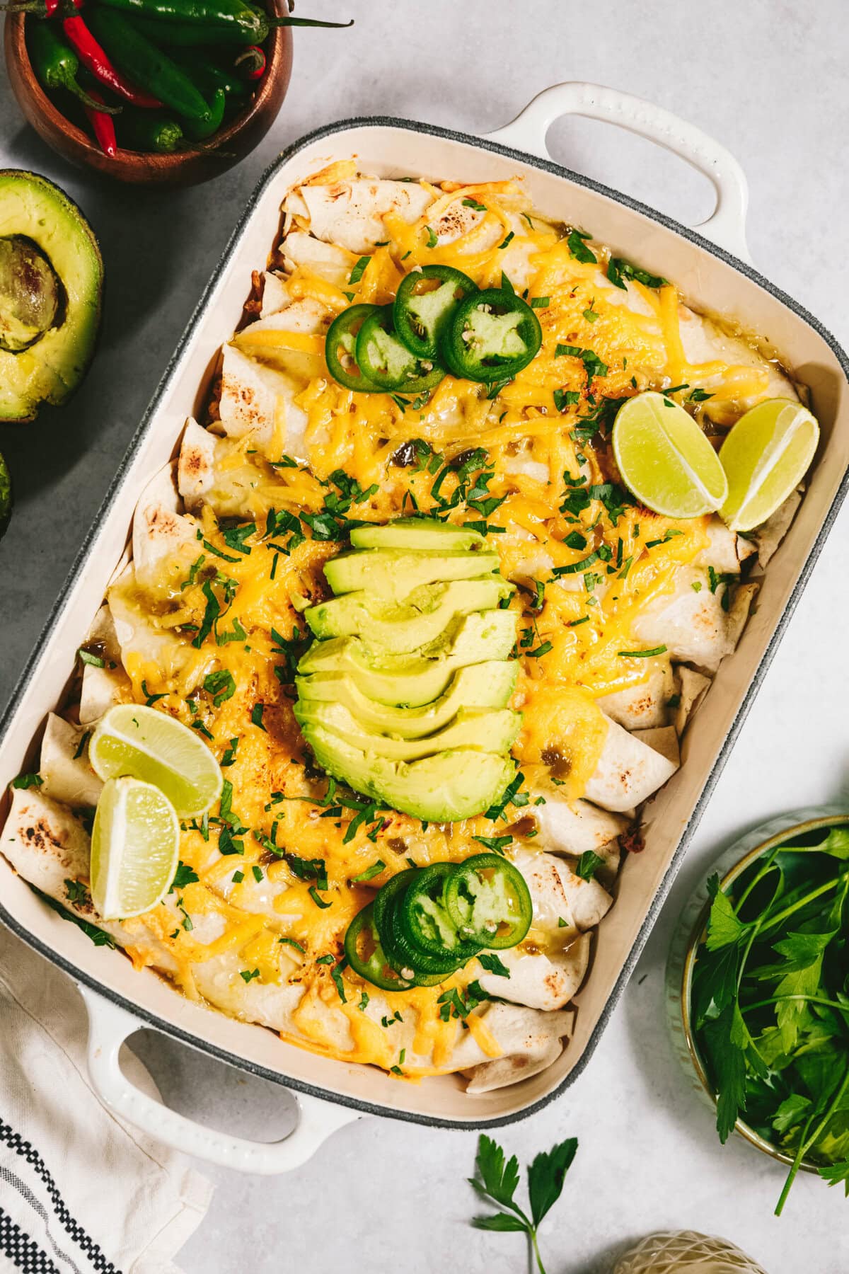 A baking dish filled with enchiladas, topped with melted cheese, sliced avocados, jalapeños, cilantro, and lime wedges. Surrounding the dish are fresh avocado, herbs, and chili peppers.