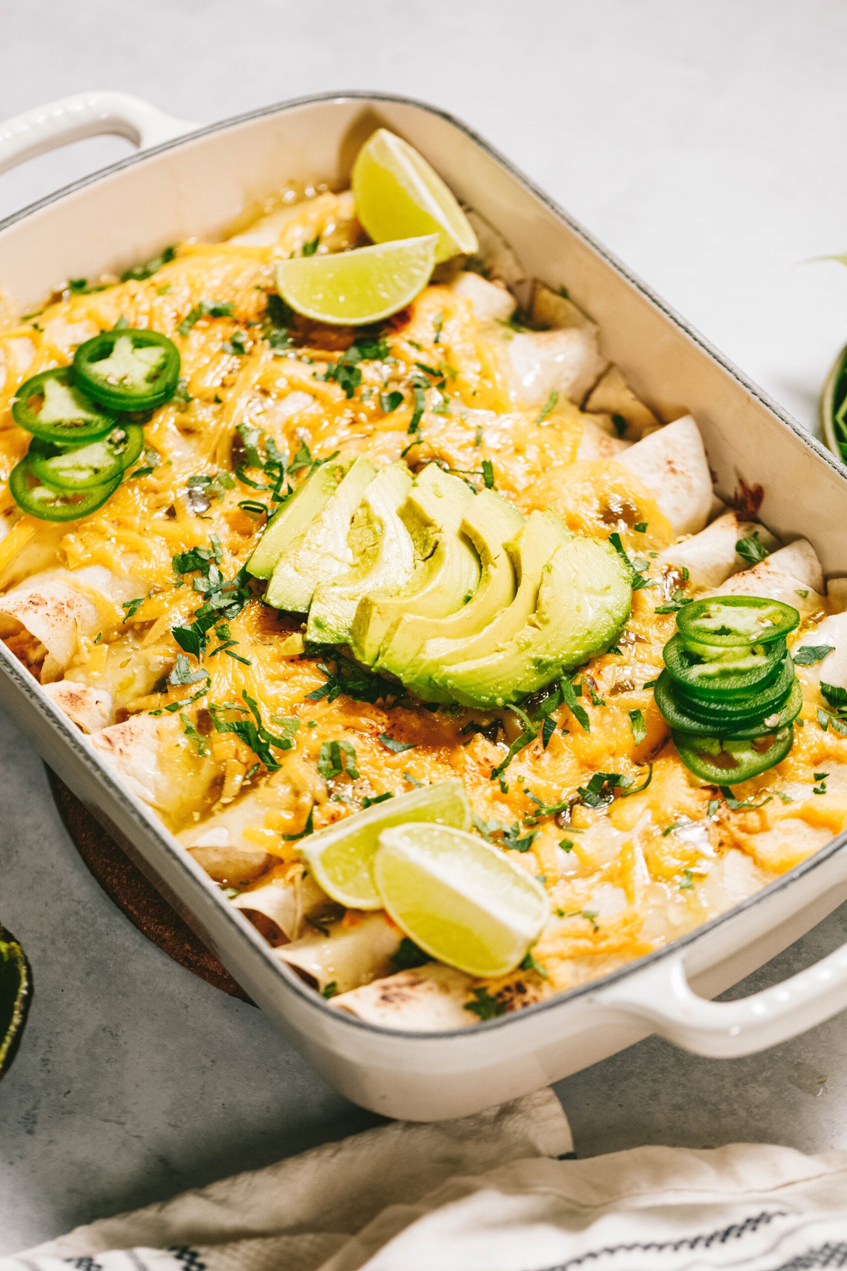 A baking dish filled with enchiladas topped with melted cheese, sliced ​​avocado, lime wedges, and jalapeño slices on a light-colored surface.