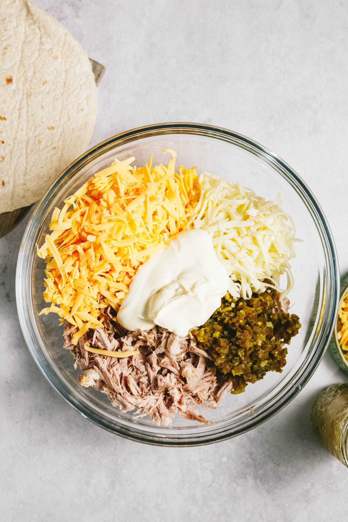 A glass bowl containing shredded cheese, mayonnaise, shredded meat, and diced pickles. A tortilla and a jar of additional ingredients are beside the bowl on a white surface.