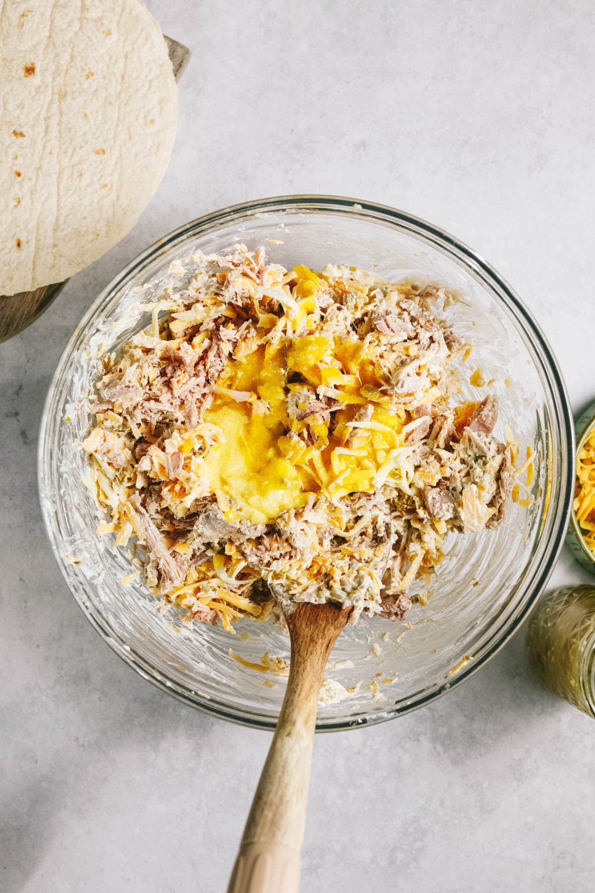 A bowl full of ingredients including shredded meat, cheese, and yellow sauce. There is a wooden spoon in the bowl. Nearby is a tortilla and a jar of grated cheese.