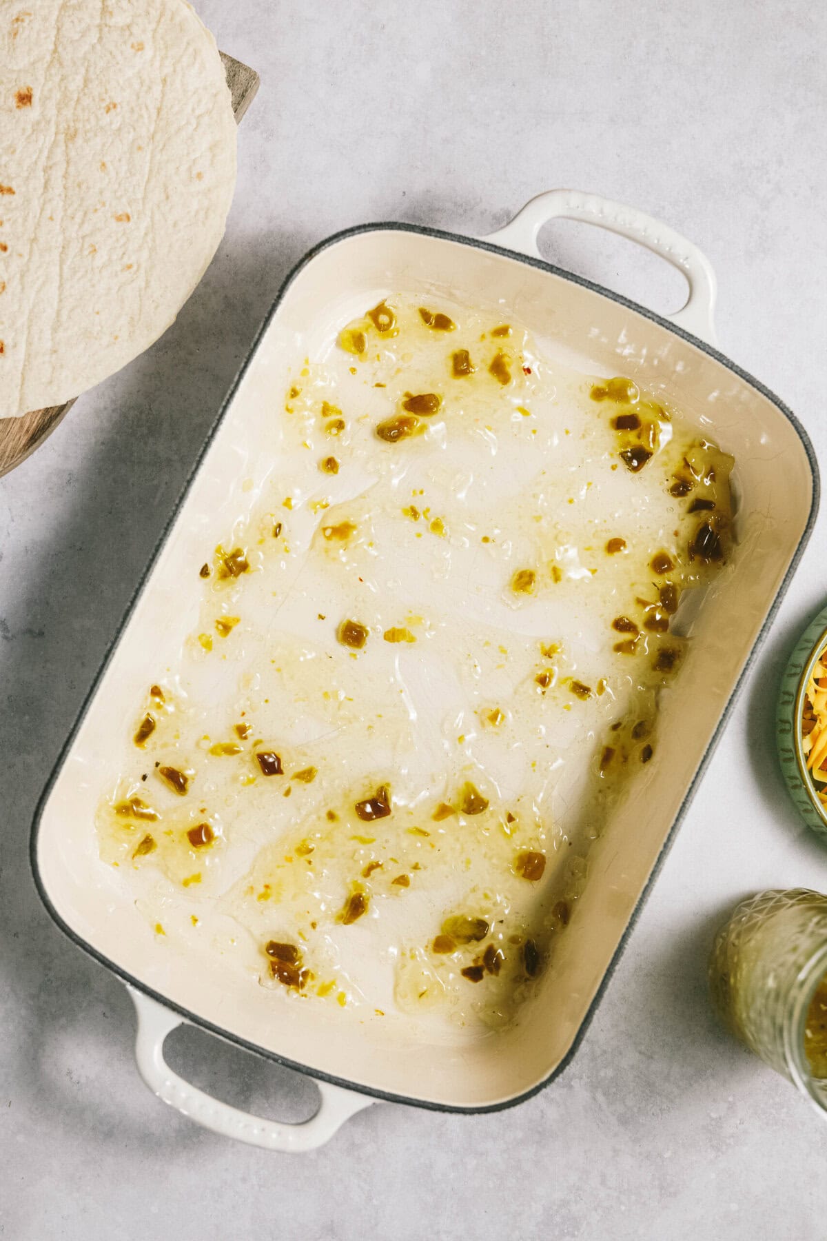 A baking dish with melted butter and garlic, a partially visible tortilla, a plate of grated cheese and a jar of salsa verde on a marble countertop.