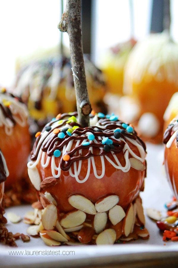 A gourmet caramel apple coated with caramel, nuts, and drizzled chocolate, topped with colorful sprinkles and white icing, on a light background.