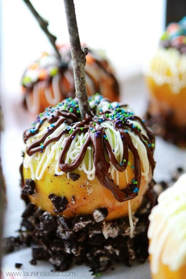 Close-up of a gourmet caramel apple coated with crushed cookies and drizzled with white and dark chocolate, topped with colorful sprinkles.