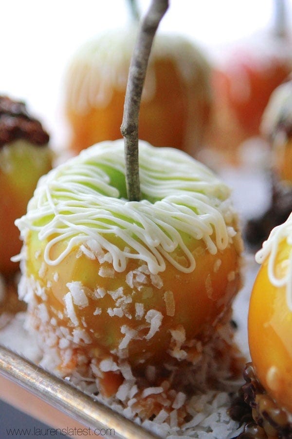 A close-up of a gourmet caramel apple with a light green coating, drizzled with white chocolate, and rolled in shredded coconut, displayed on a tray.