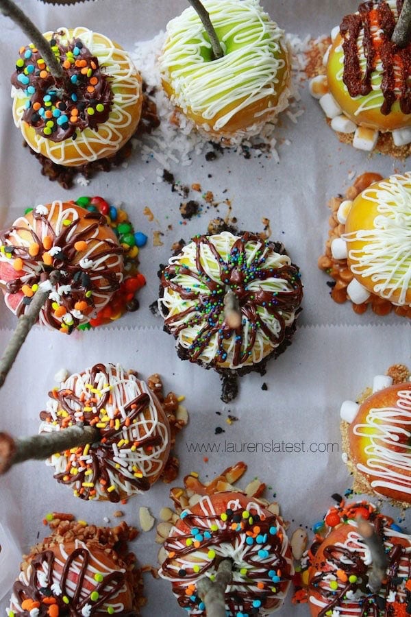 A tray of gourmet caramel apples, each topped with colorful sprinkles, drizzles of chocolate and white icing, and assorted candies. Sticks are inserted into the apples as handles.