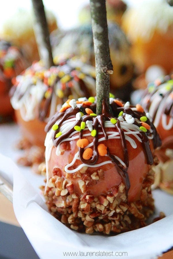 Close-up of a gourmet caramel apple covered in chopped nuts, drizzled with chocolate, and topped with colorful sprinkles, displayed on a parchment-lined surface. Other similarly decorated gourmet apples are in the background.