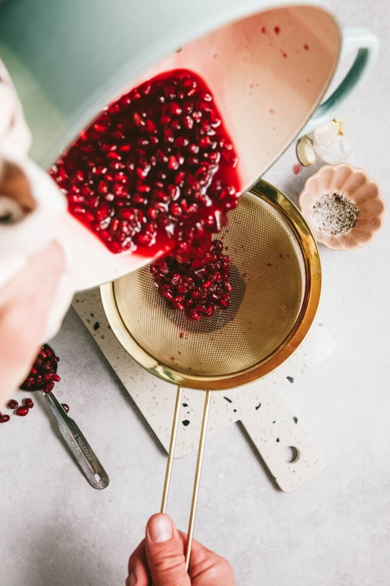 A person pouring pomegranate seeds and juice through a gold fine-mesh strainer over a bowl, with some kitchen items like spices and utensils nearby.