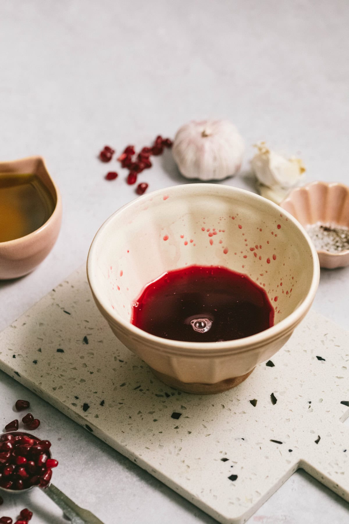 A ceramic bowl containing a red liquid sits on a speckled cutting board, surrounded by pomegranate seeds, garlic cloves, salt, pepper, and a small bowl of olive oil.