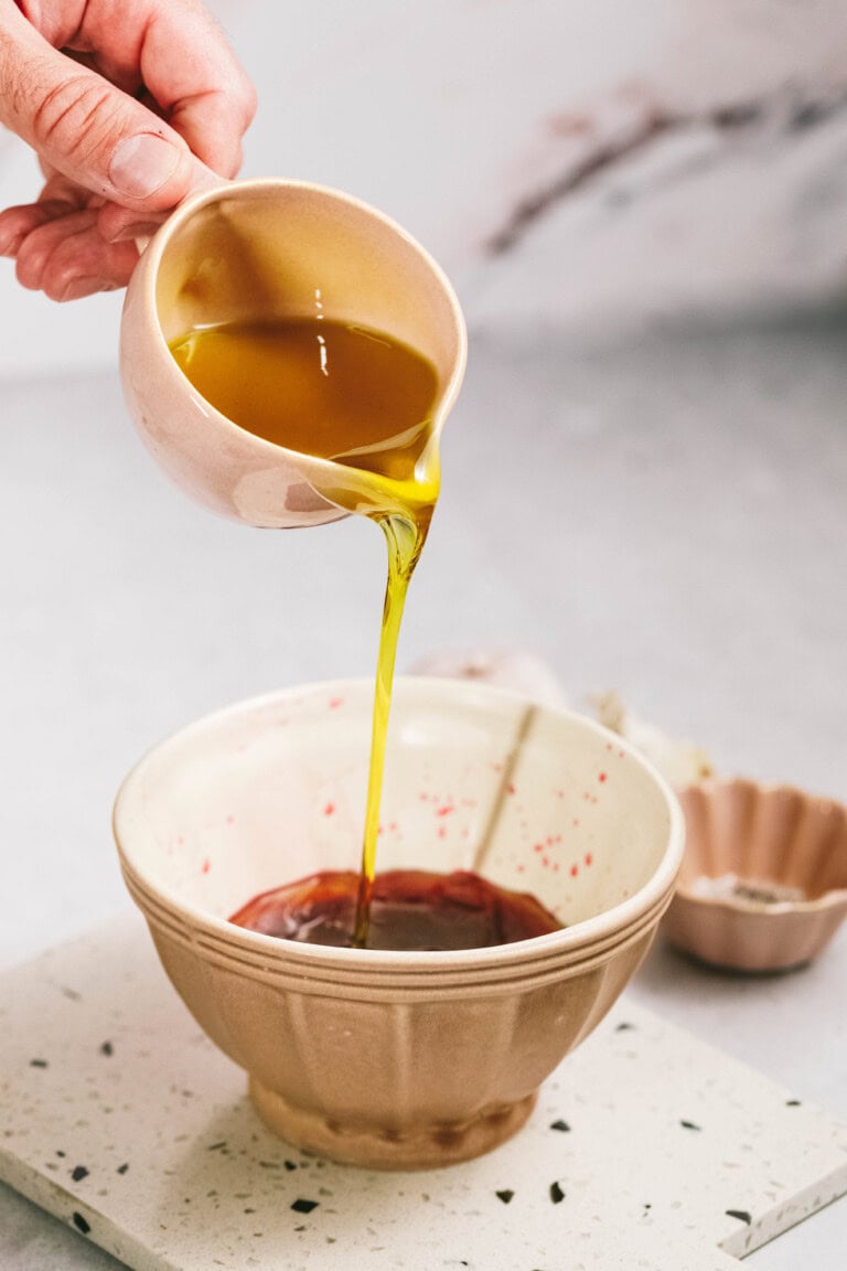 A hand pours a stream of olive oil from a small pink cup into a pink mixing bowl containing a dark liquid, possibly soy sauce, against a light-colored background.