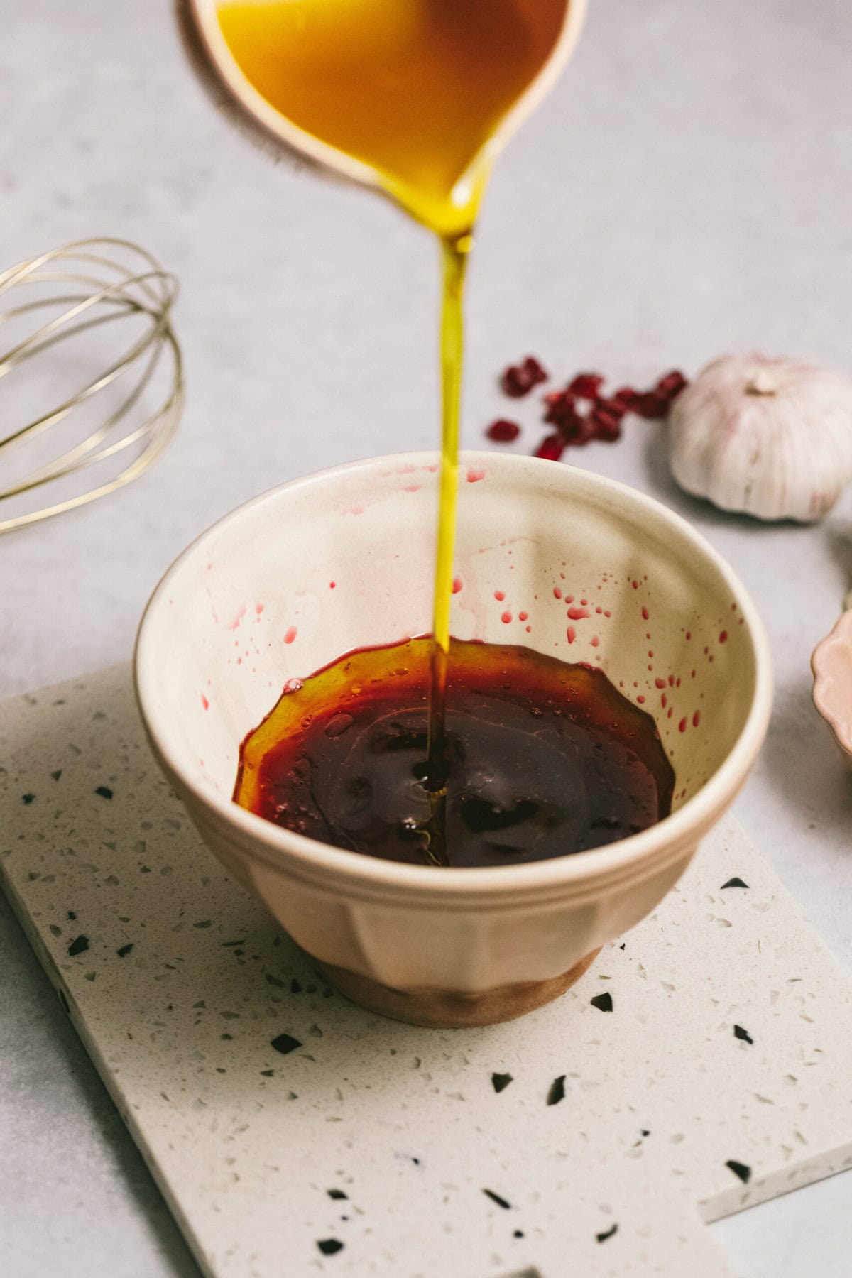 A bowl with a dark liquid being poured into it from a container. A whisk, garlic, and red berries are on the table beside it.