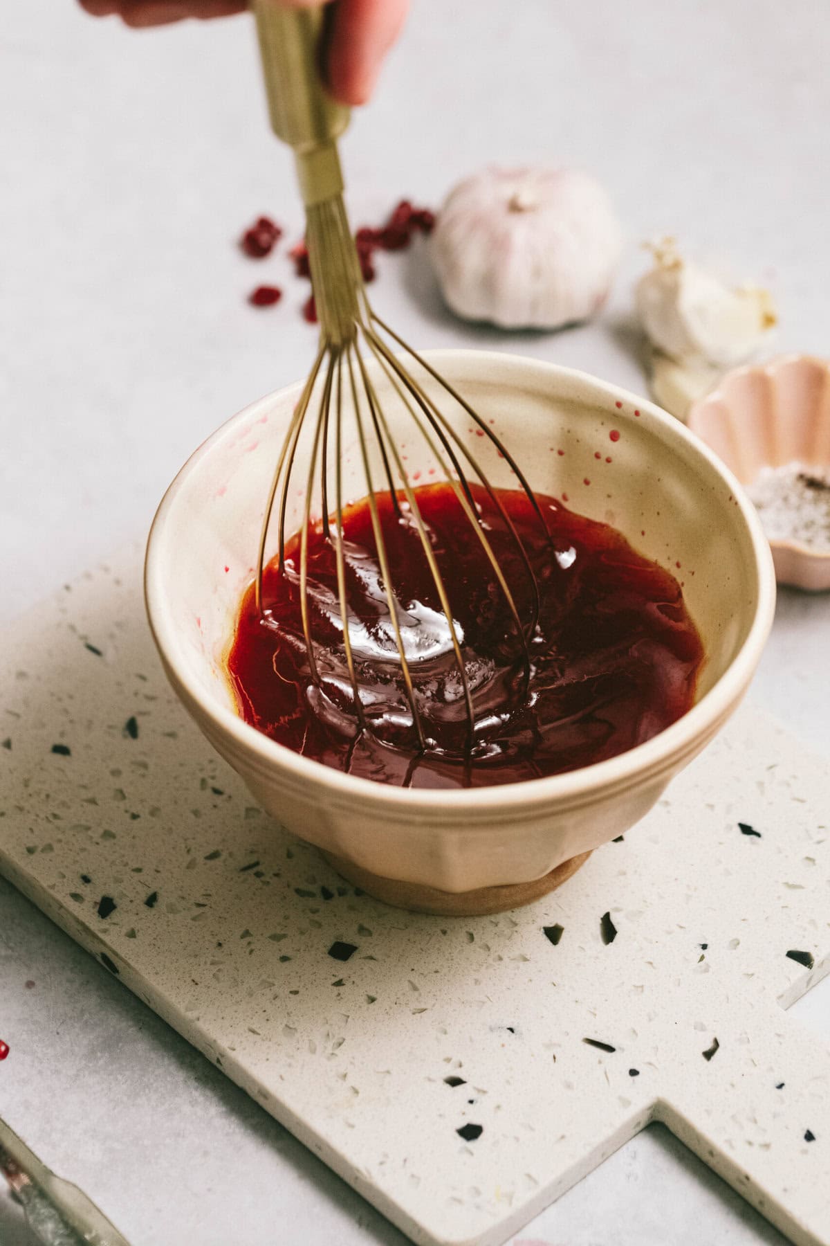 A hand holding a whisk above a bowl of red liquid on a speckled cutting board, with garlic and a small bowl of salt in the background.