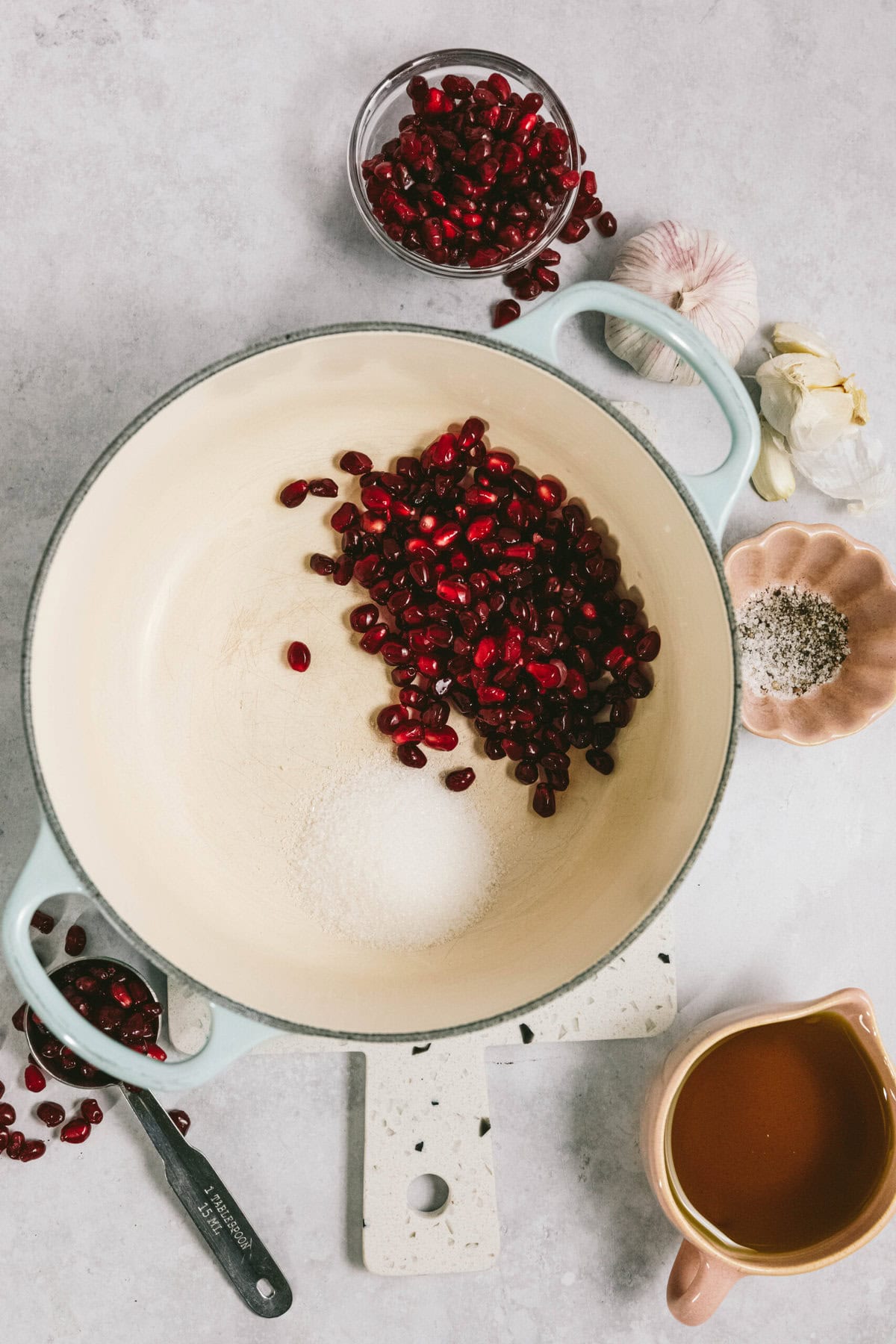 A white pot containing pomegranate seeds and sugar on a countertop, surrounded by a bowl of pomegranate seeds, garlic, spices, and a jug of liquid.