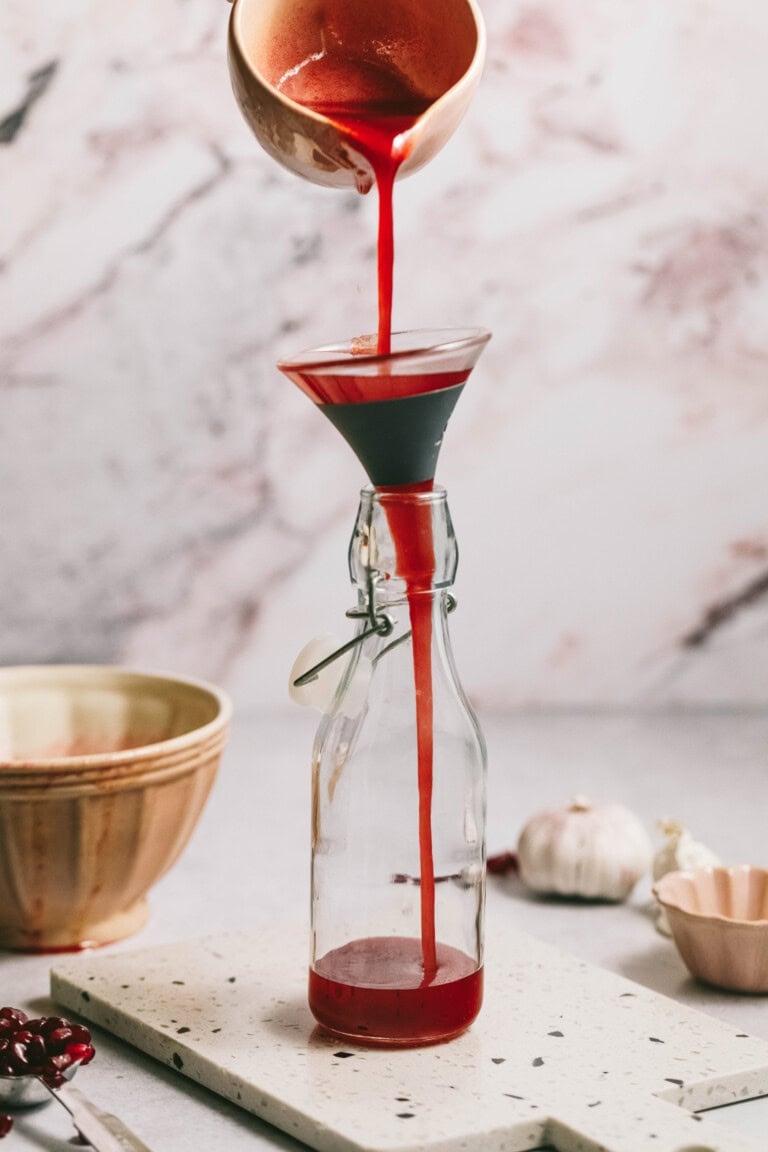 A person pours a red liquid from a bowl through a funnel into a glass bottle. The bottle is on a white surface, with a bowl and garlic in the background.