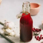 A glass bottle filled with red liquid, sealed with a clasp, sits on a speckled cutting board near garlic cloves and pomegranate seeds.