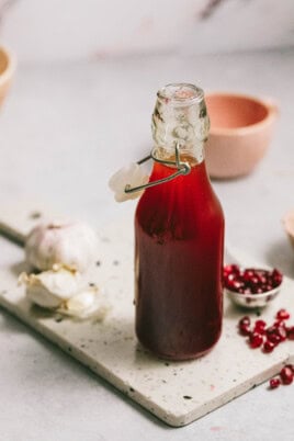 A glass bottle filled with red liquid, sealed with a clasp, sits on a speckled cutting board near garlic cloves and pomegranate seeds.