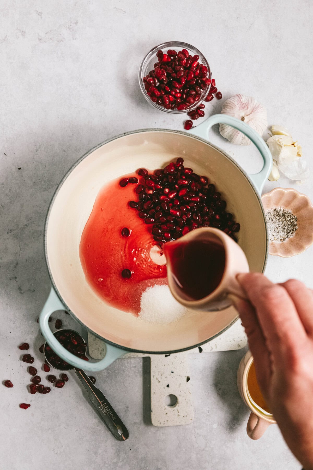 Hand pouring juice into a pot with pomegranate seeds and sugar, surrounded by bowls containing pomegranate seeds, spices, garlic, and a measuring spoon.