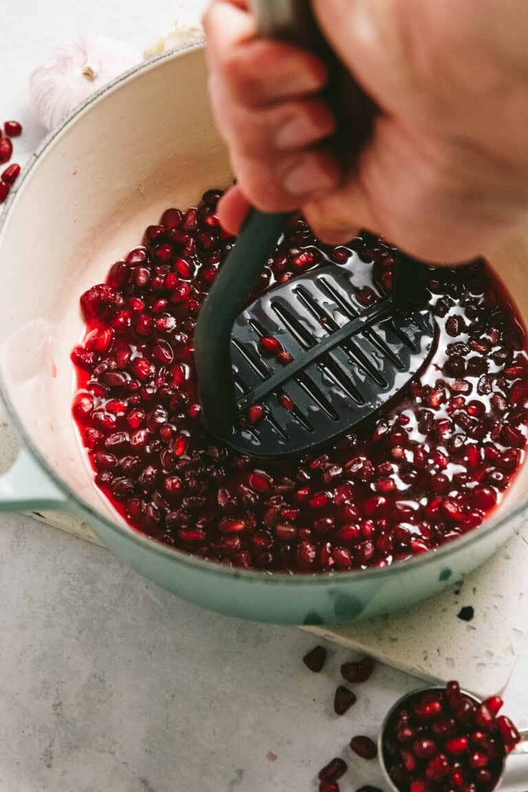 A person mashes pomegranate seeds in a pot with a potato masher, surrounded by scattered seeds and cloves of garlic.