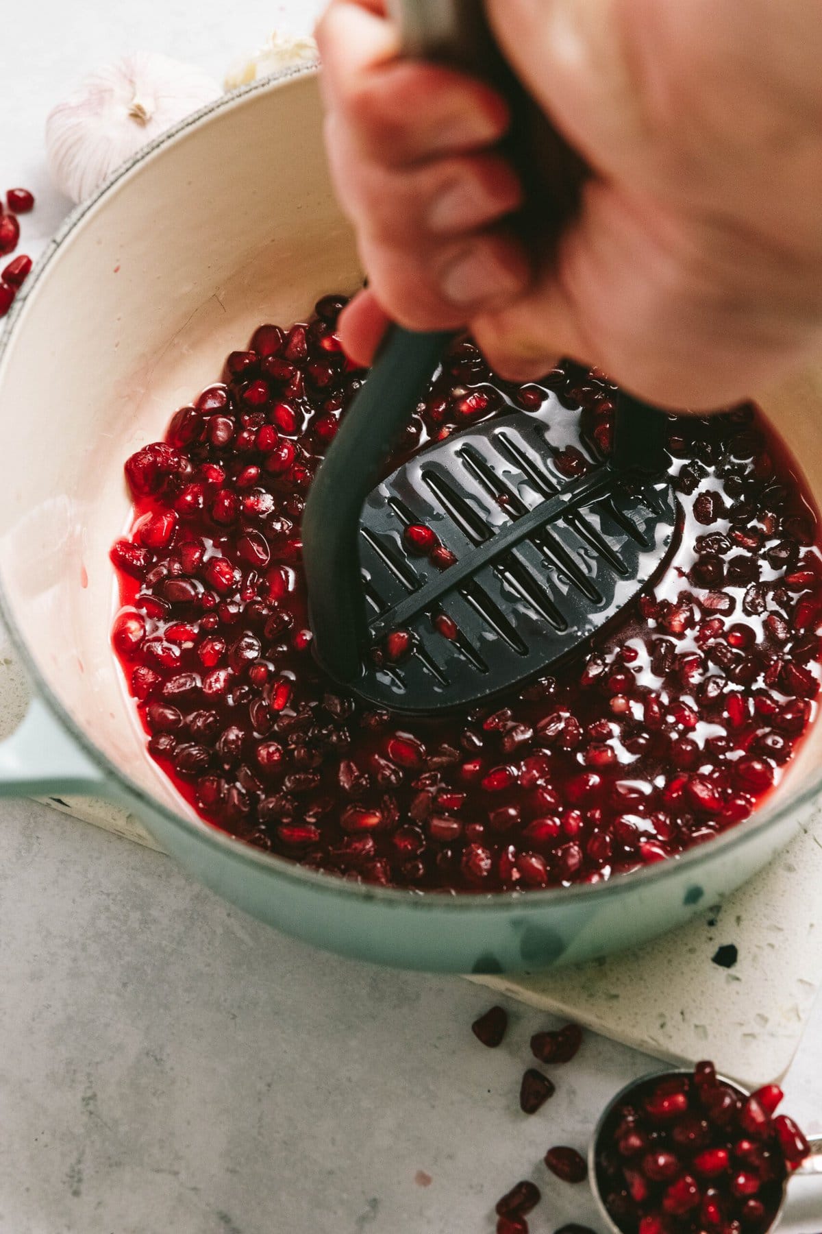 A person mashes pomegranate seeds in a pot with a potato masher, surrounded by scattered seeds and cloves of garlic.