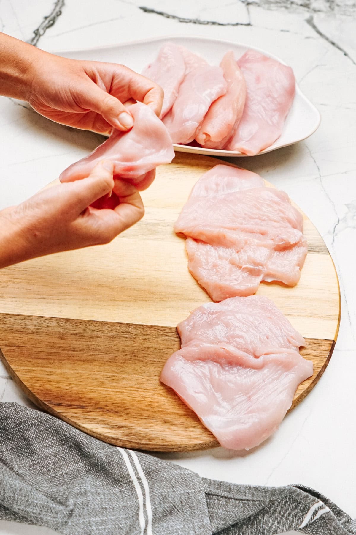 Hands preparing raw chicken fillets on a wooden board, with more fillets on a white plate in the background.