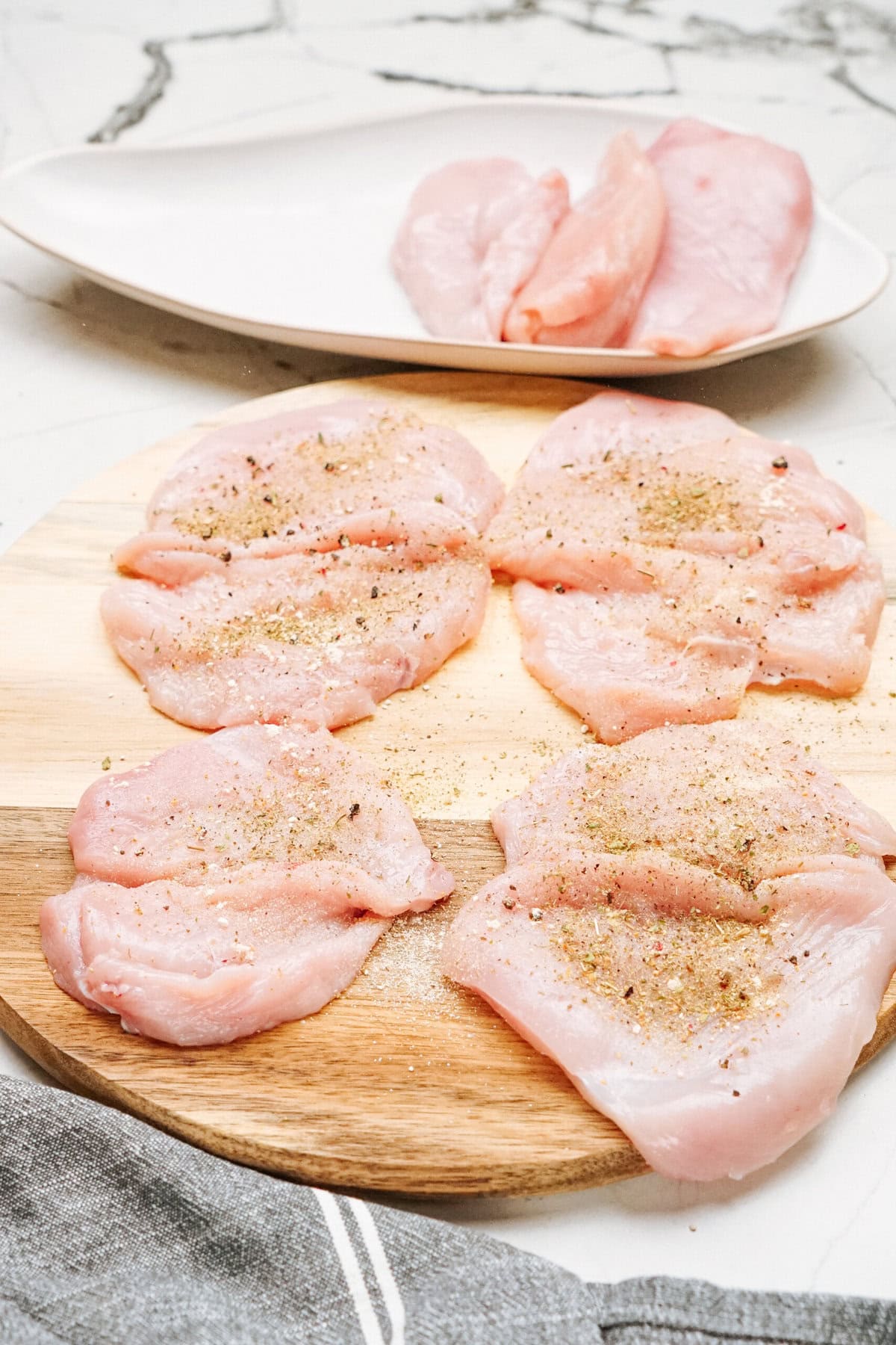 Raw chicken cutlets seasoned with spices on a wooden cutting board, with more cutlets on a white plate in the background.