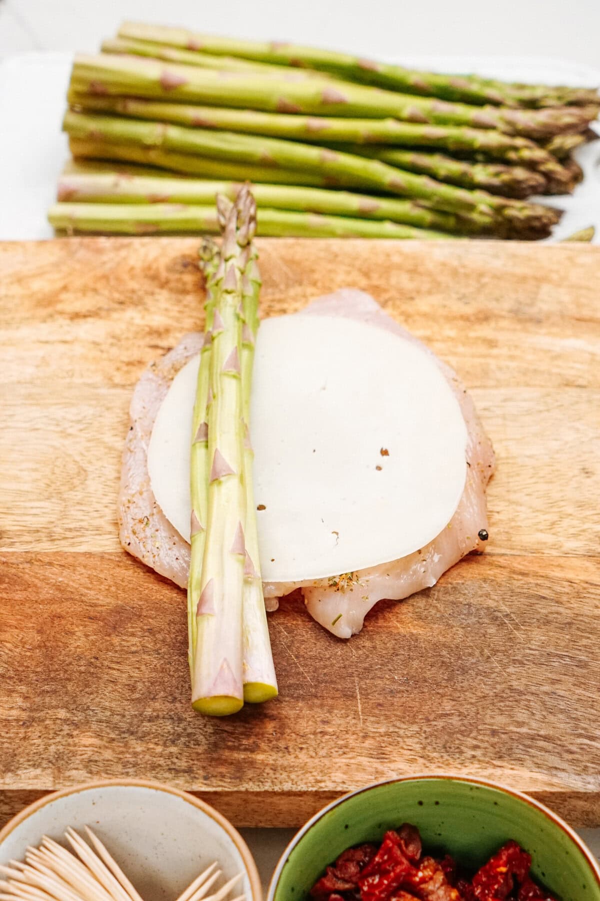 Chicken breast topped with cheese and asparagus spears on a wooden cutting board. Bowls of sundried tomatoes and toothpicks are nearby. Fresh asparagus in the background.