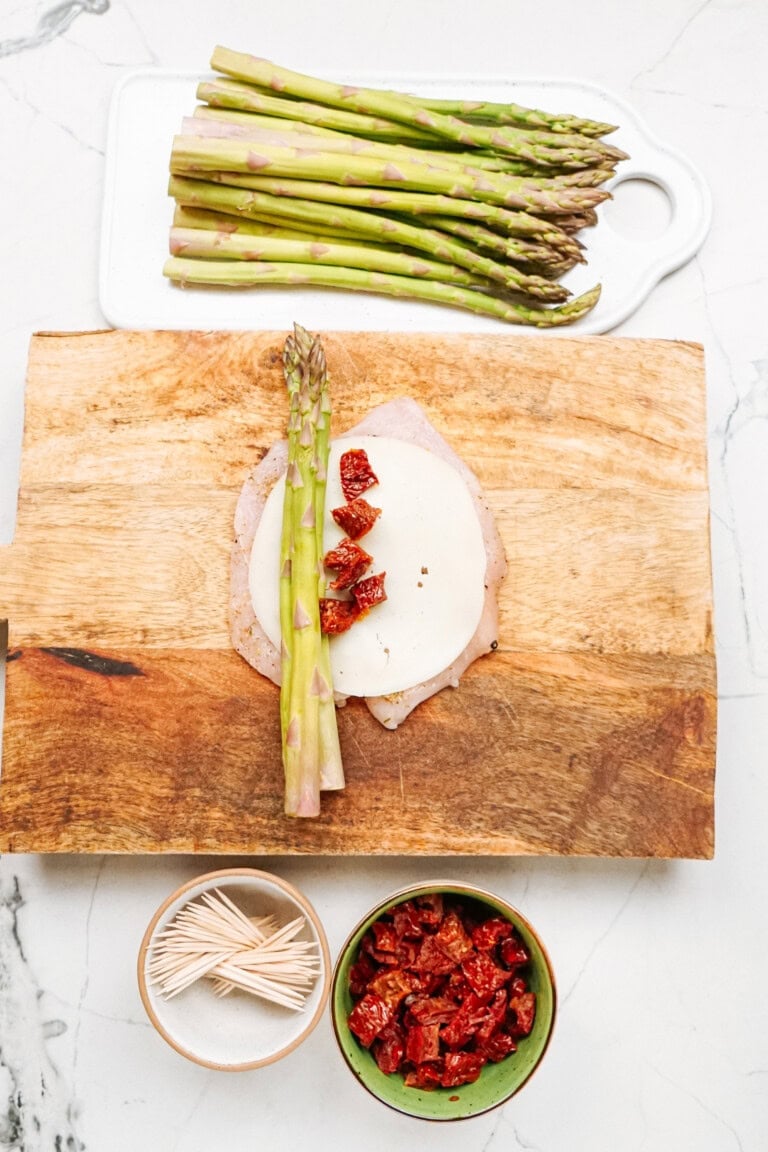 Asparagus spears, cheese slice, and turkey slice on a wooden board, surrounded by bowls of sun-dried tomatoes and toothpicks, with extra asparagus on a white cutting board.