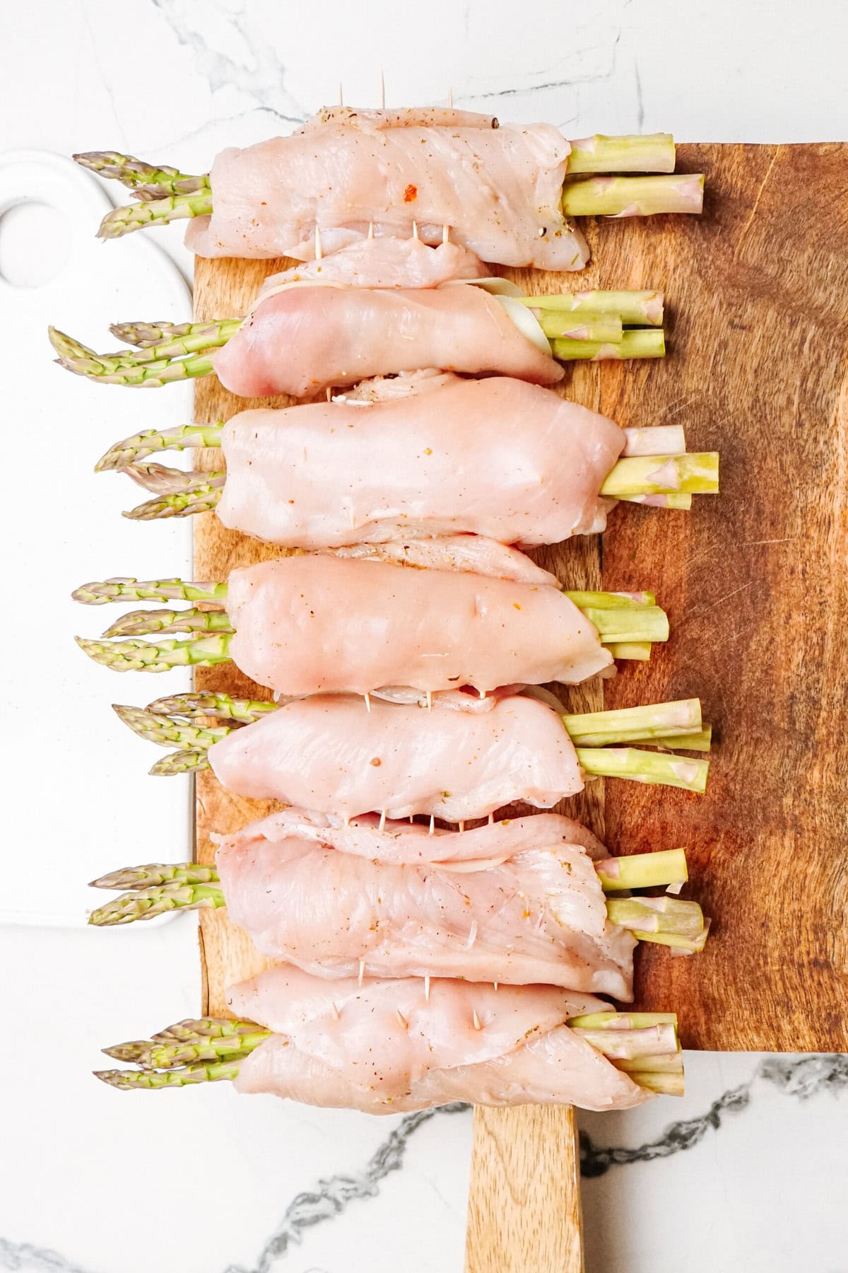 Raw chicken breast fillets wrapped around asparagus spears, secured with toothpicks, arranged neatly on a wooden cutting board.