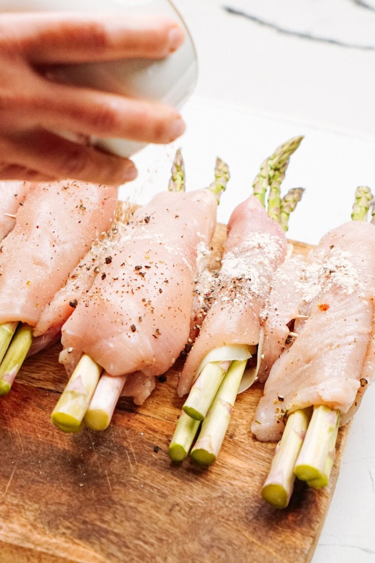 Chicken fillets wrapped around asparagus spears are being seasoned on a wooden board.