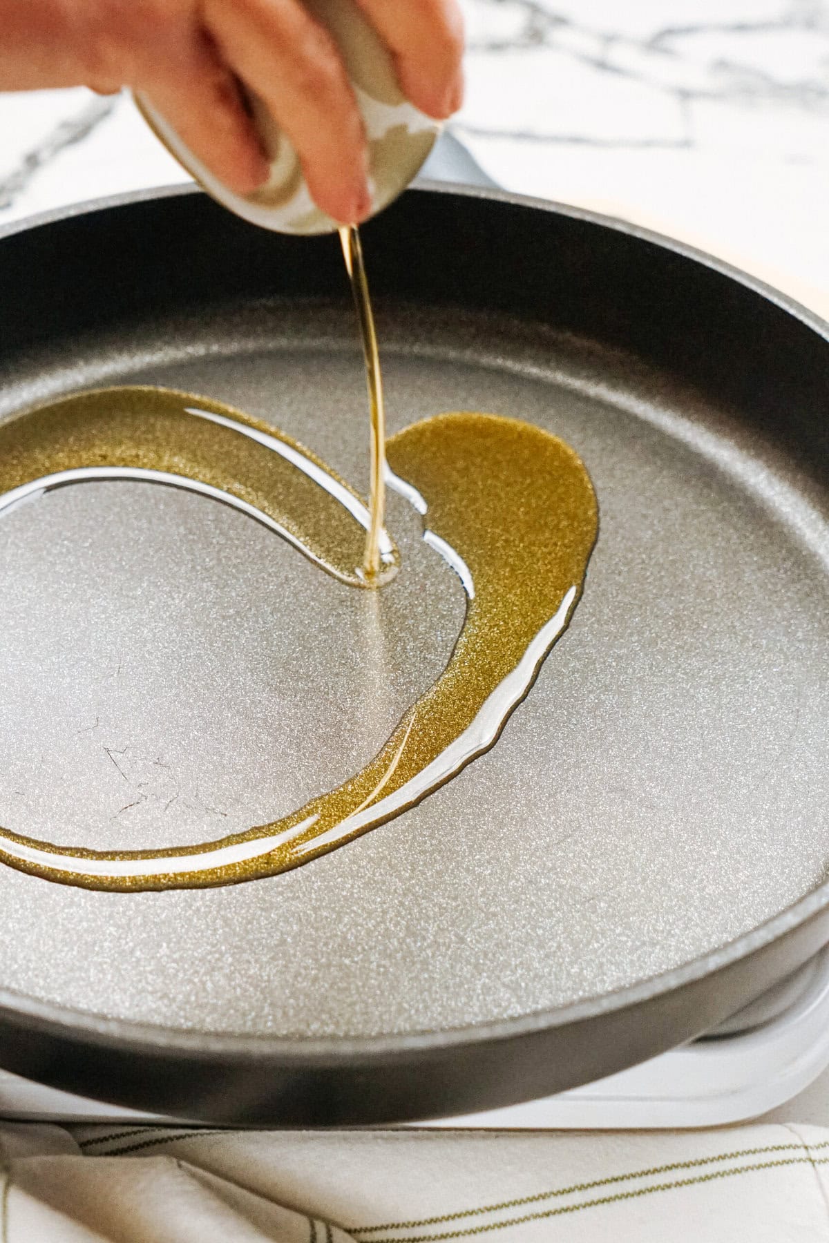 A hand pours oil from a small container into a frying pan on a stovetop.