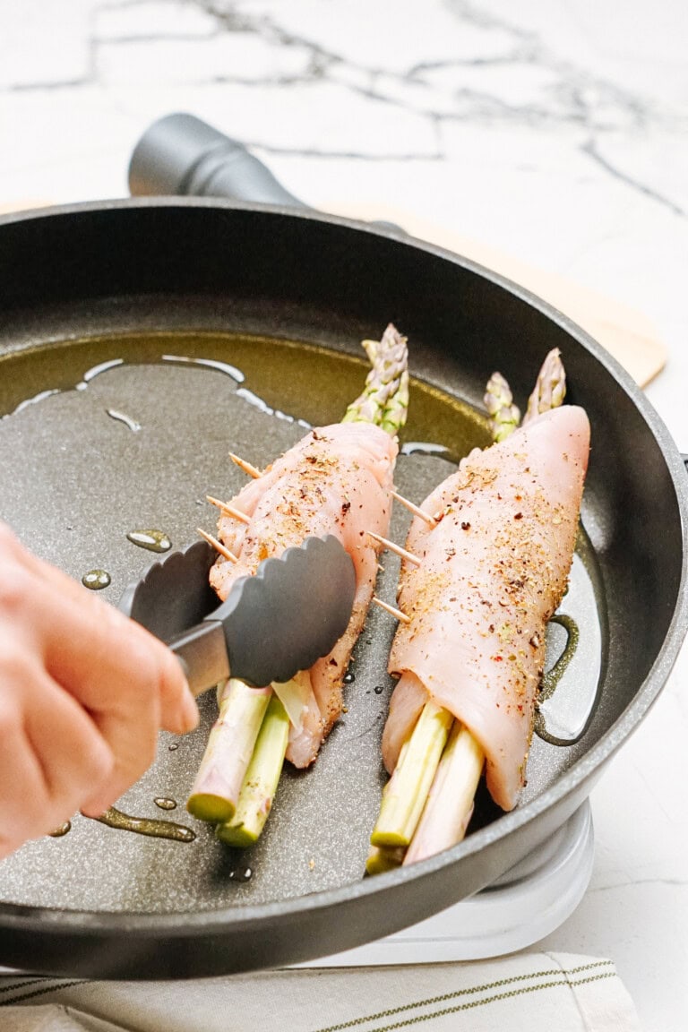 Two chicken breasts wrapped around asparagus spears being cooked in a pan, secured with toothpicks, and seasoned with pepper. A hand holding tongs is seen at the left.