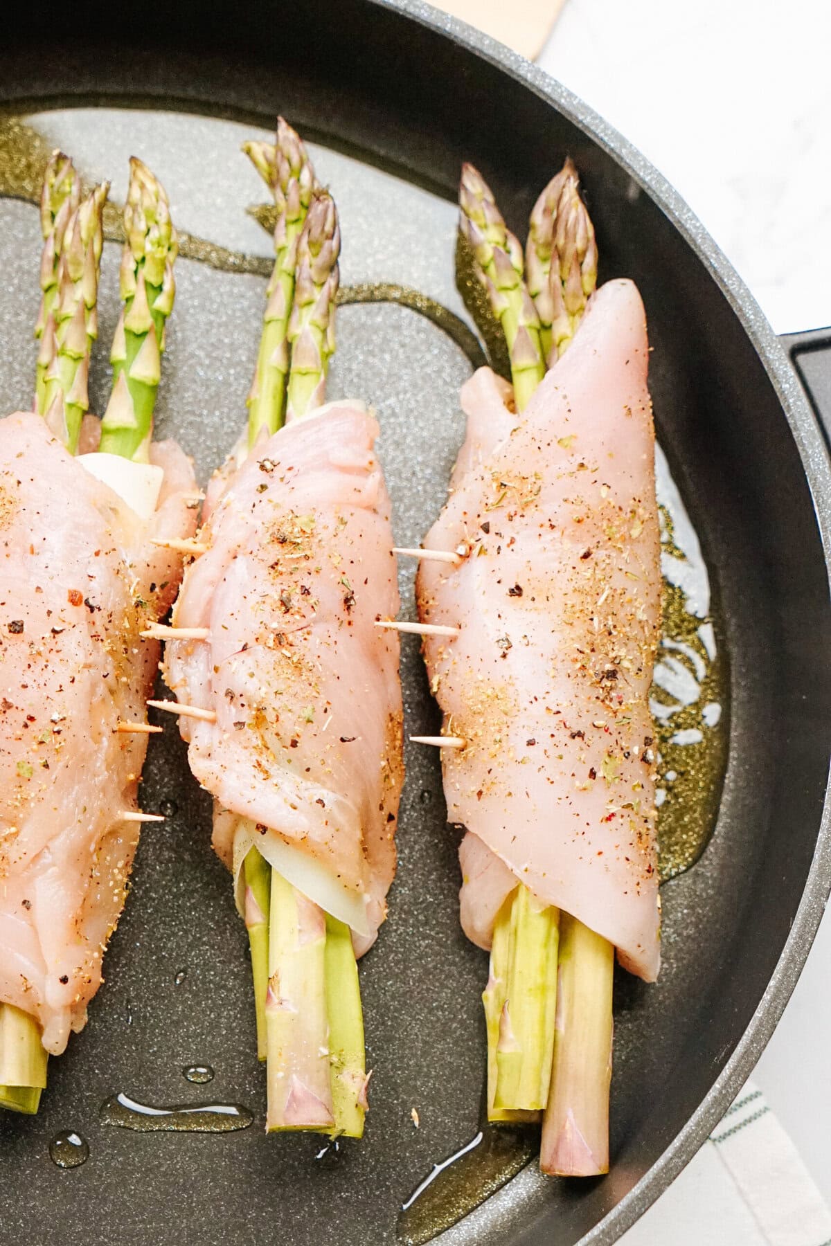 Raw chicken breasts stuffed with asparagus and secured with toothpicks in a pan, sprinkled with seasoning.