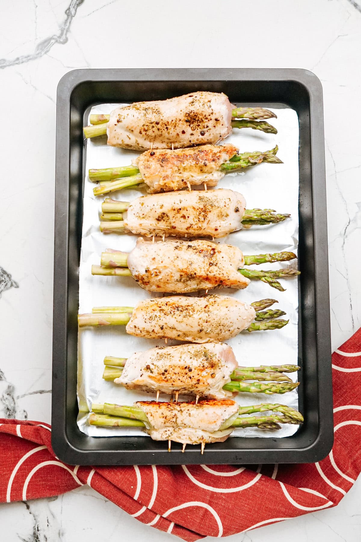 A baking tray of asparagus wrapped in seasoned chicken fillets, placed on a white marble surface next to a red patterned cloth.