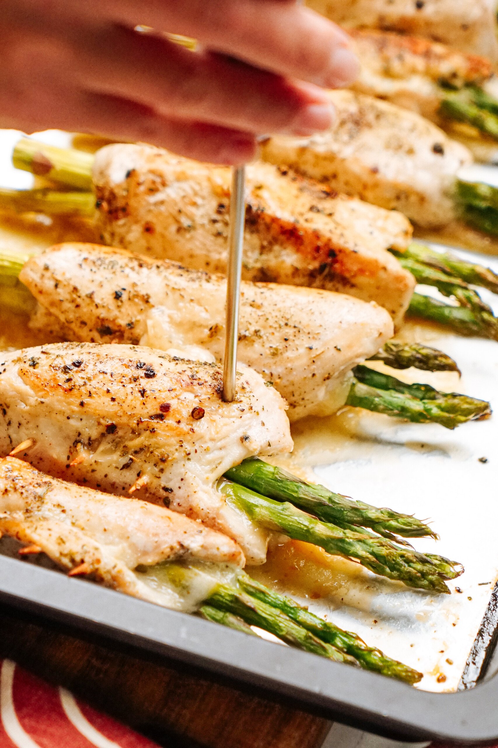 A hand holds a thermometer measuring the temperature of baked chicken breasts stuffed with asparagus on a baking tray.