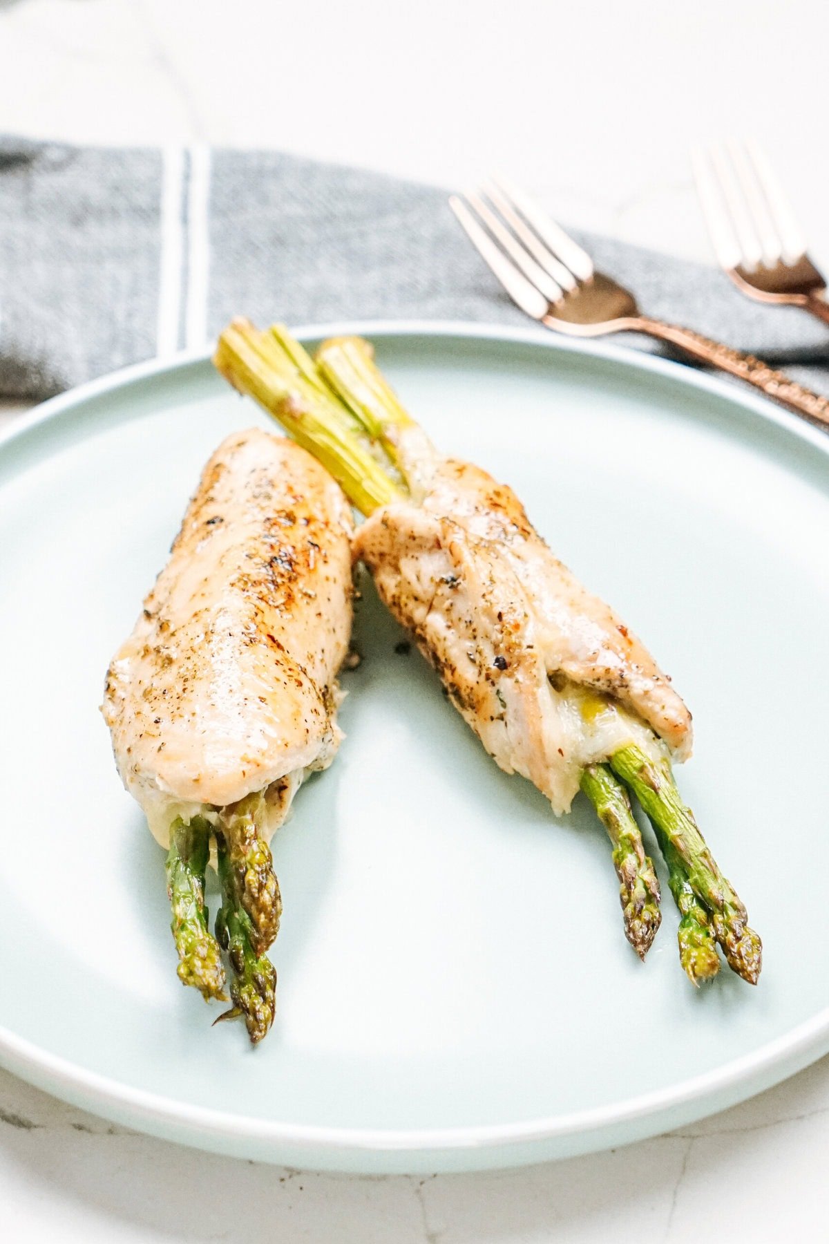 Two chicken breasts wrapped around asparagus spears, served on a light blue plate with a gray cloth and two forks in the background.