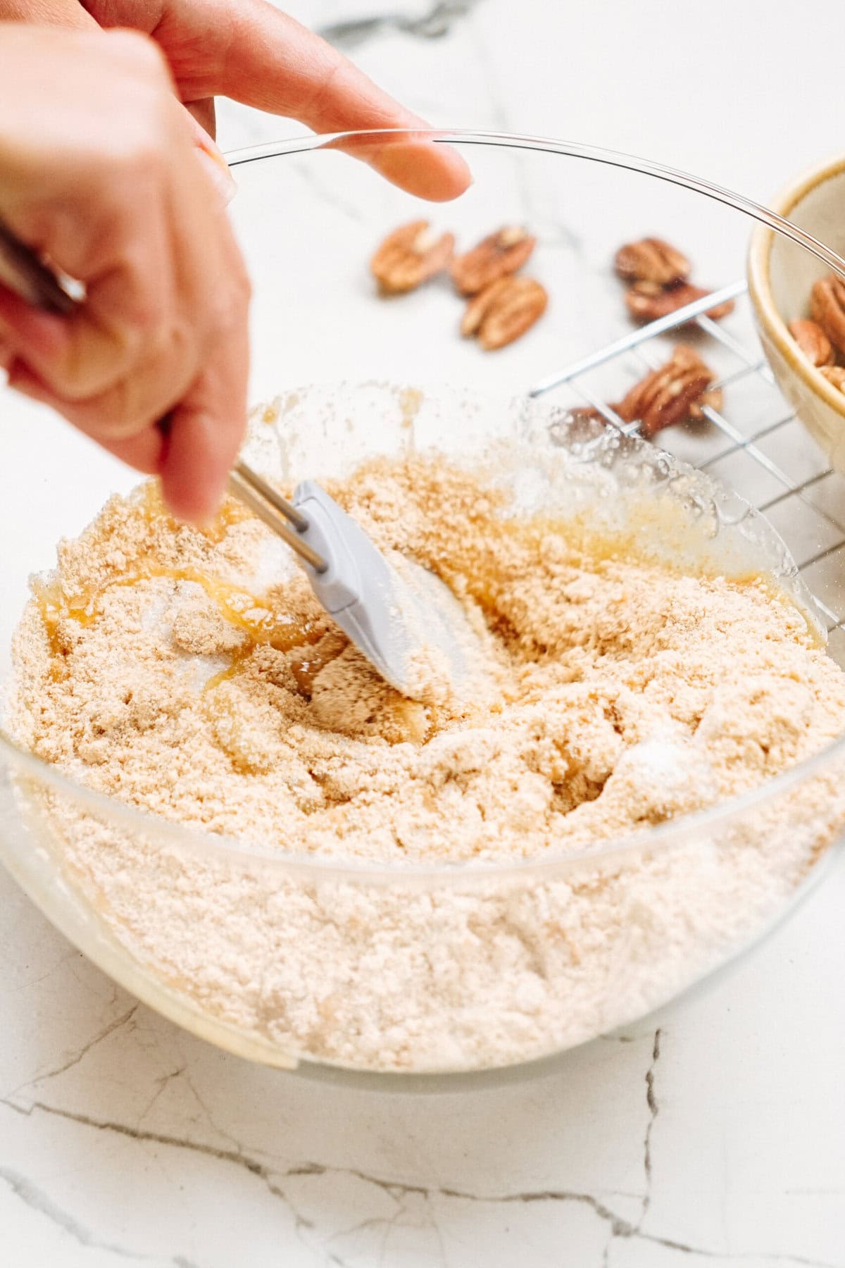 A hand using a spatula to mix dry ingredients in a glass bowl, with pecans scattered on a marble counter.