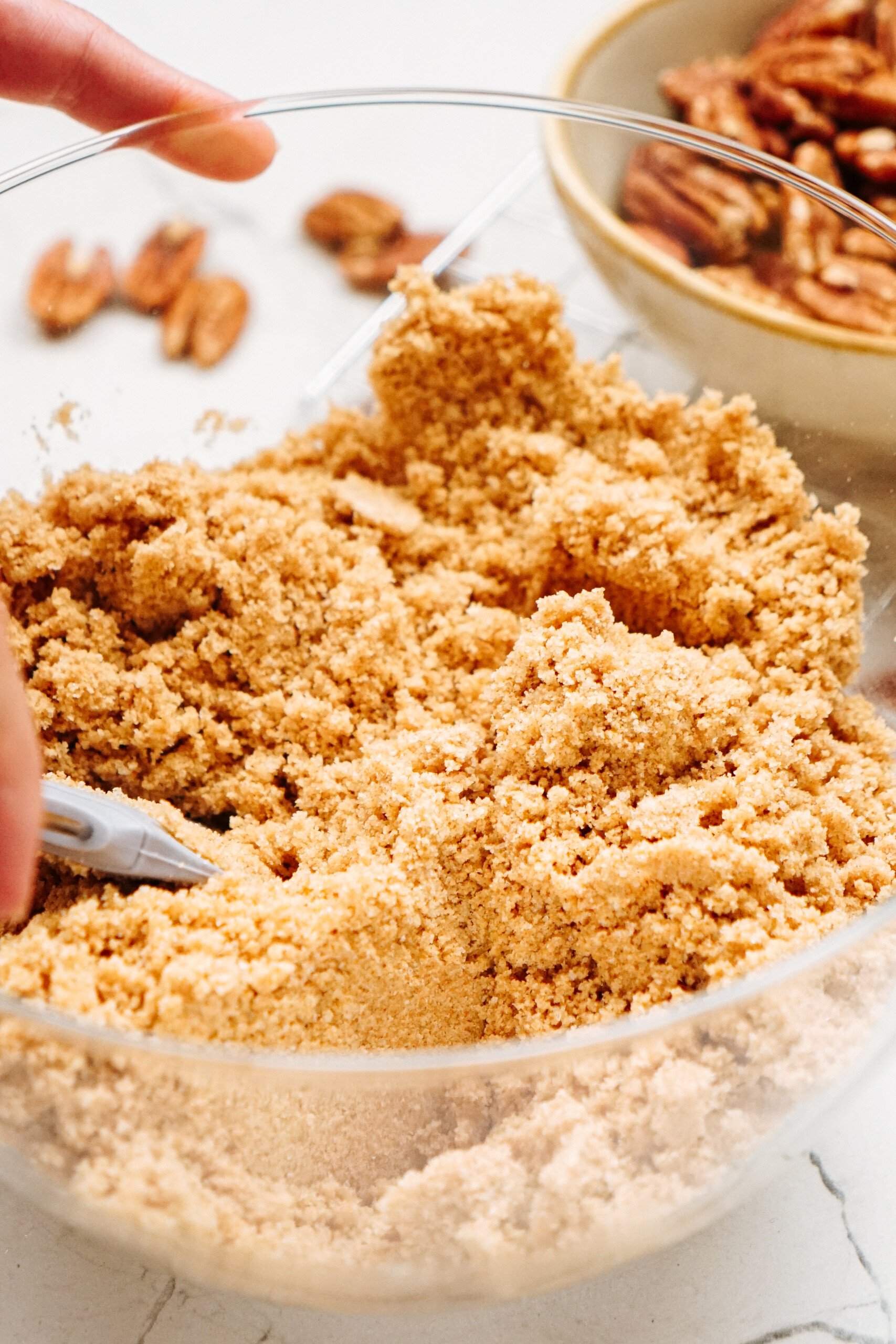 A hand mixing brown sugar in a clear bowl using a utensil. A bowl of pecans is in the background.