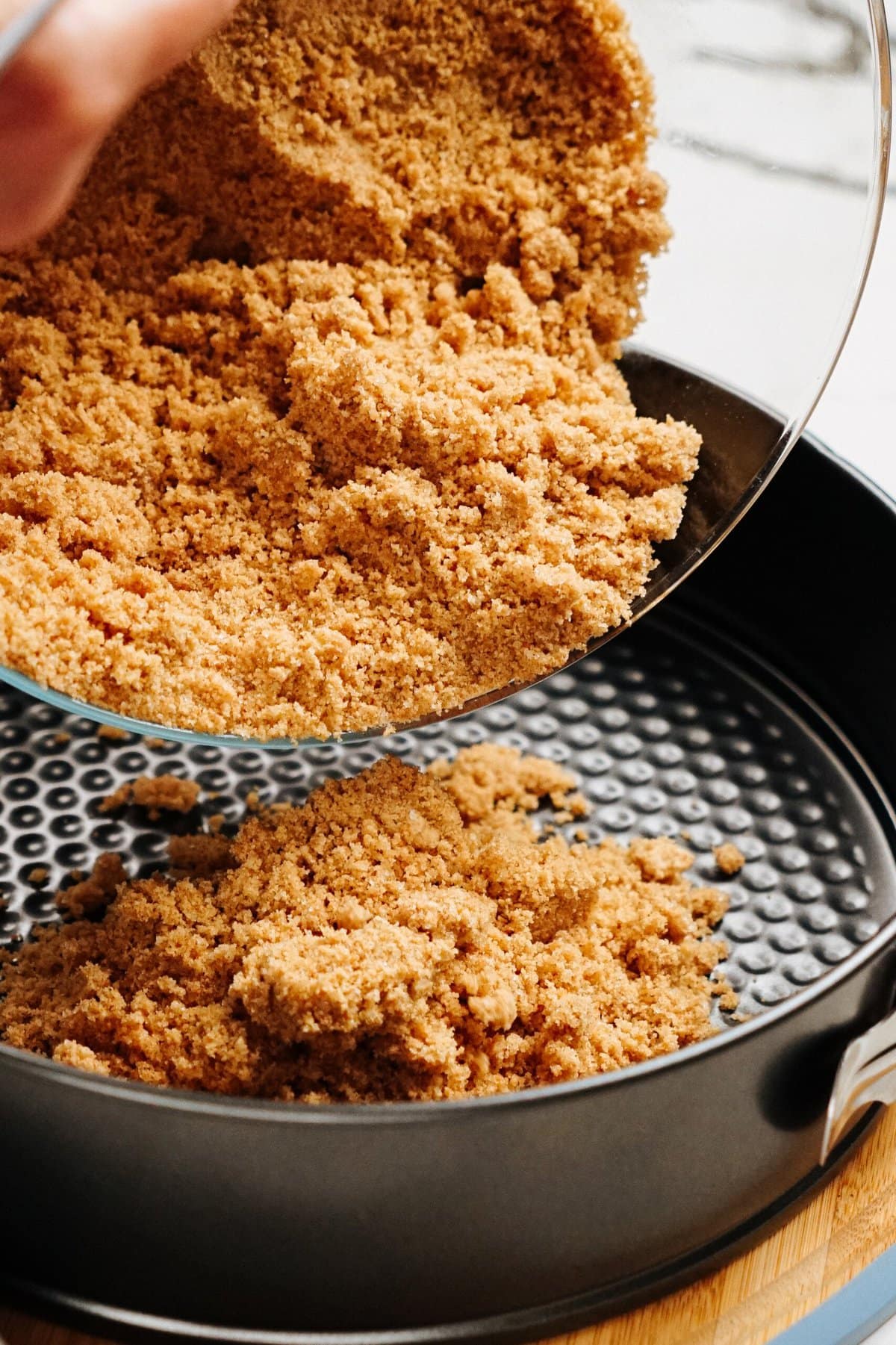 Crushed graham crackers being poured into a round springform pan on a wooden surface.