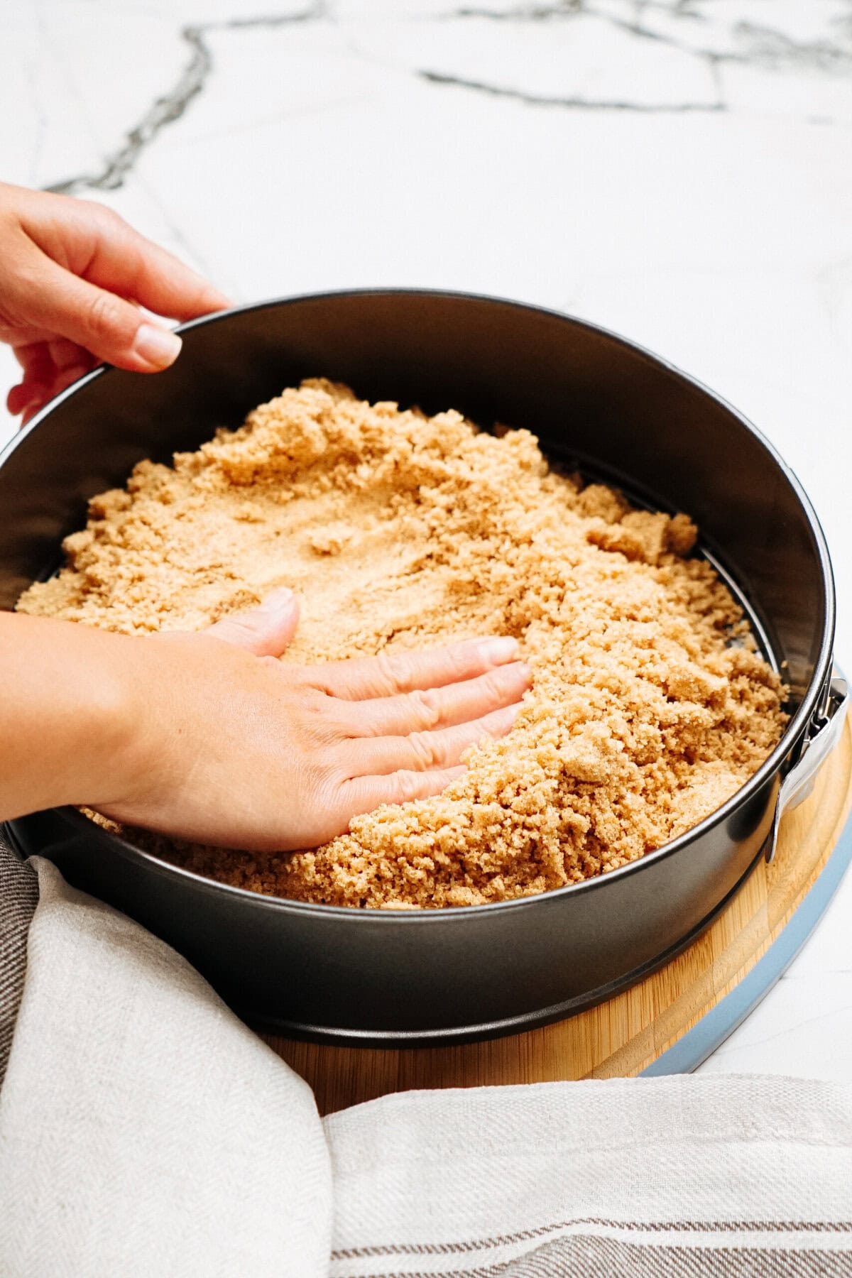 Hands pressing crumb mixture into a springform pan on a wooden board.