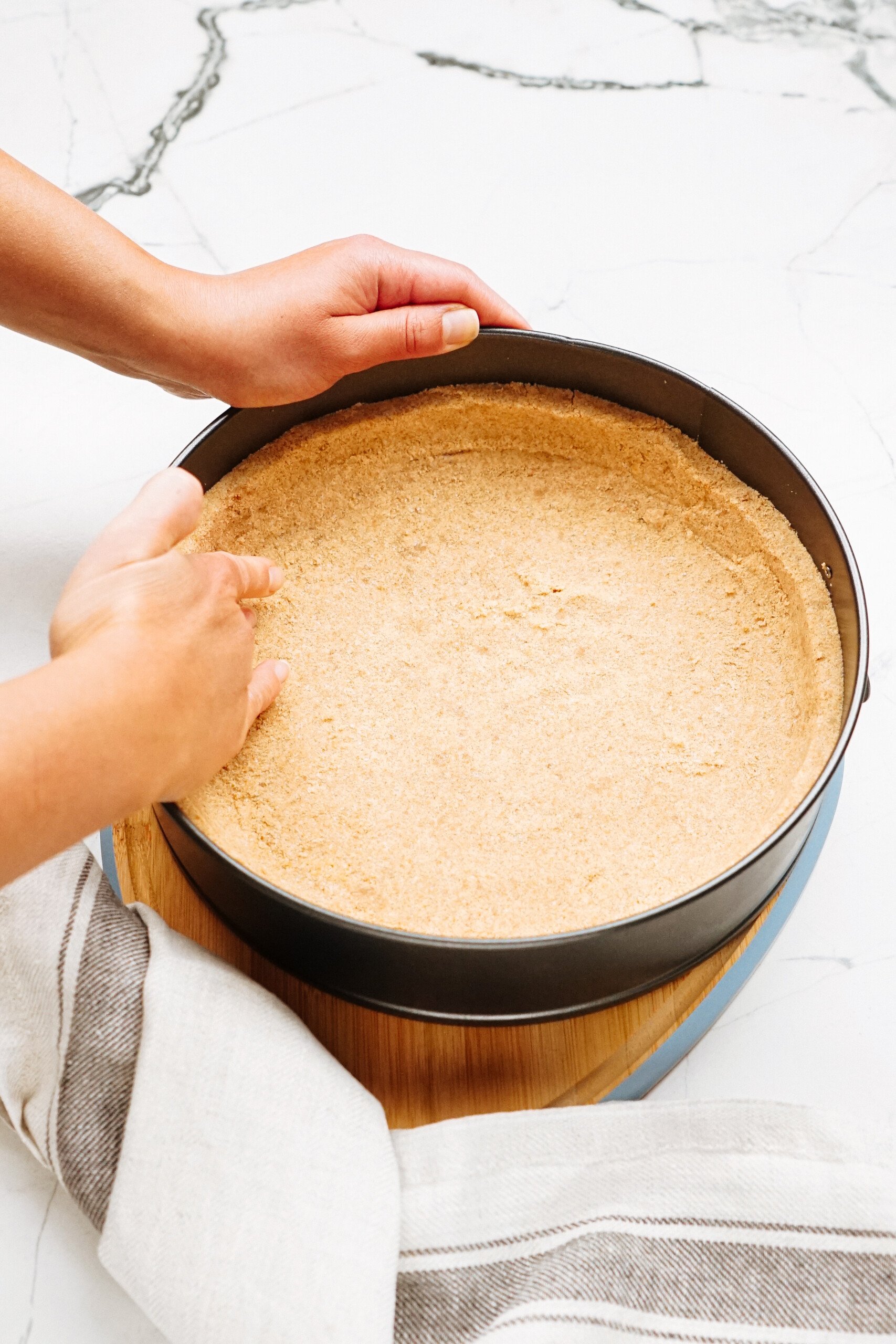 Hands pressing crumb crust into a round baking pan, on a marble surface with a striped cloth nearby.