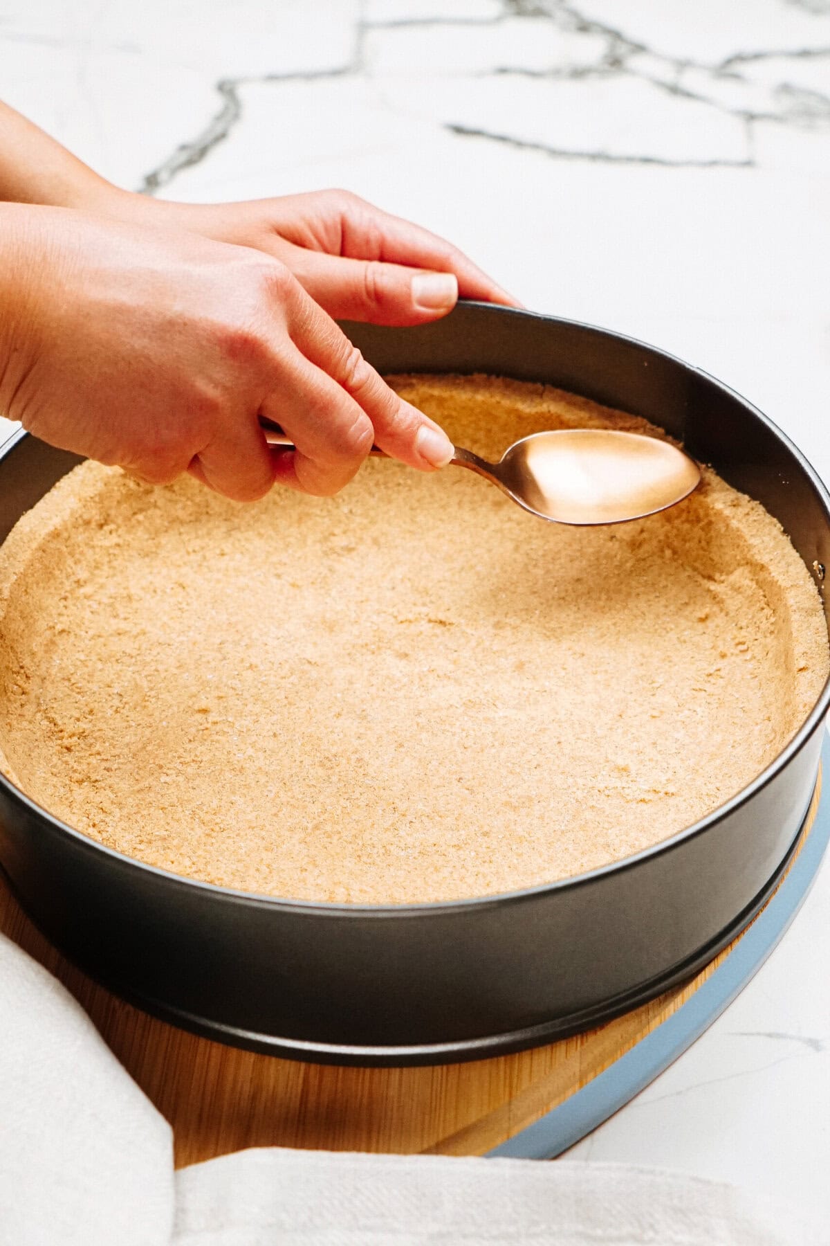 Hands pressing crumb crust into a round baking pan with a spoon on a marble countertop.