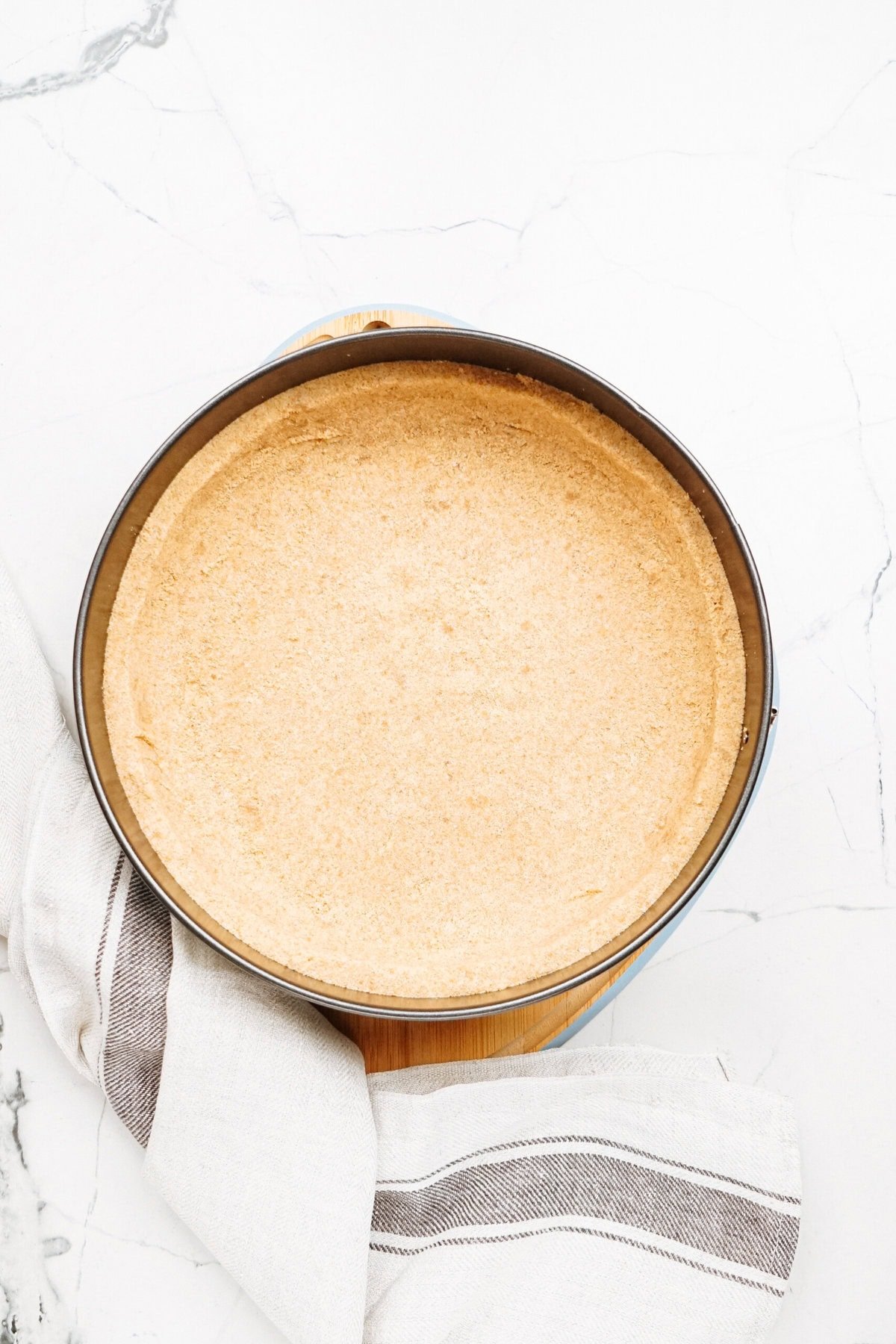 A round, unbaked graham cracker crust in a metal pan on a marble surface, with a striped kitchen towel nearby.