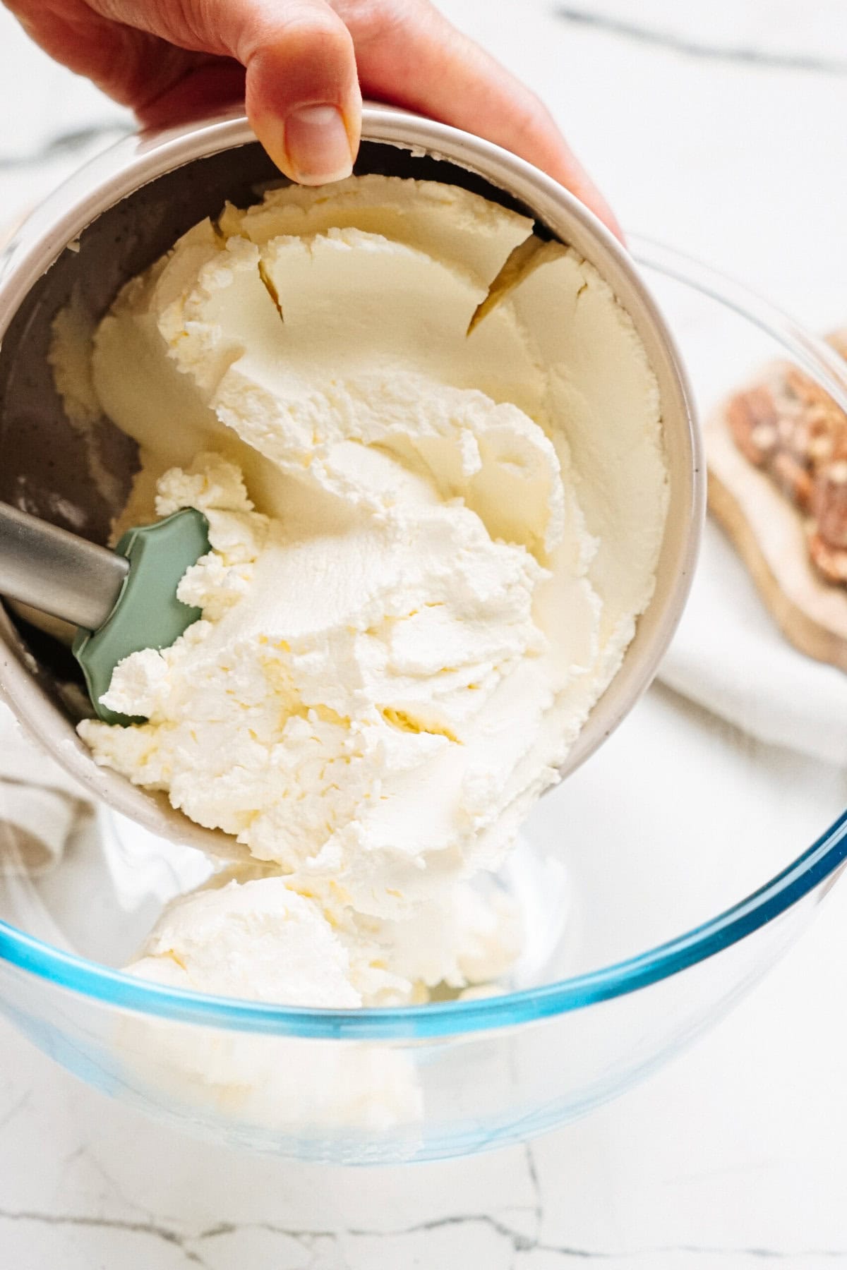 A hand holds a metal bowl, pouring white ice cream into a clear glass bowl on a marble surface.