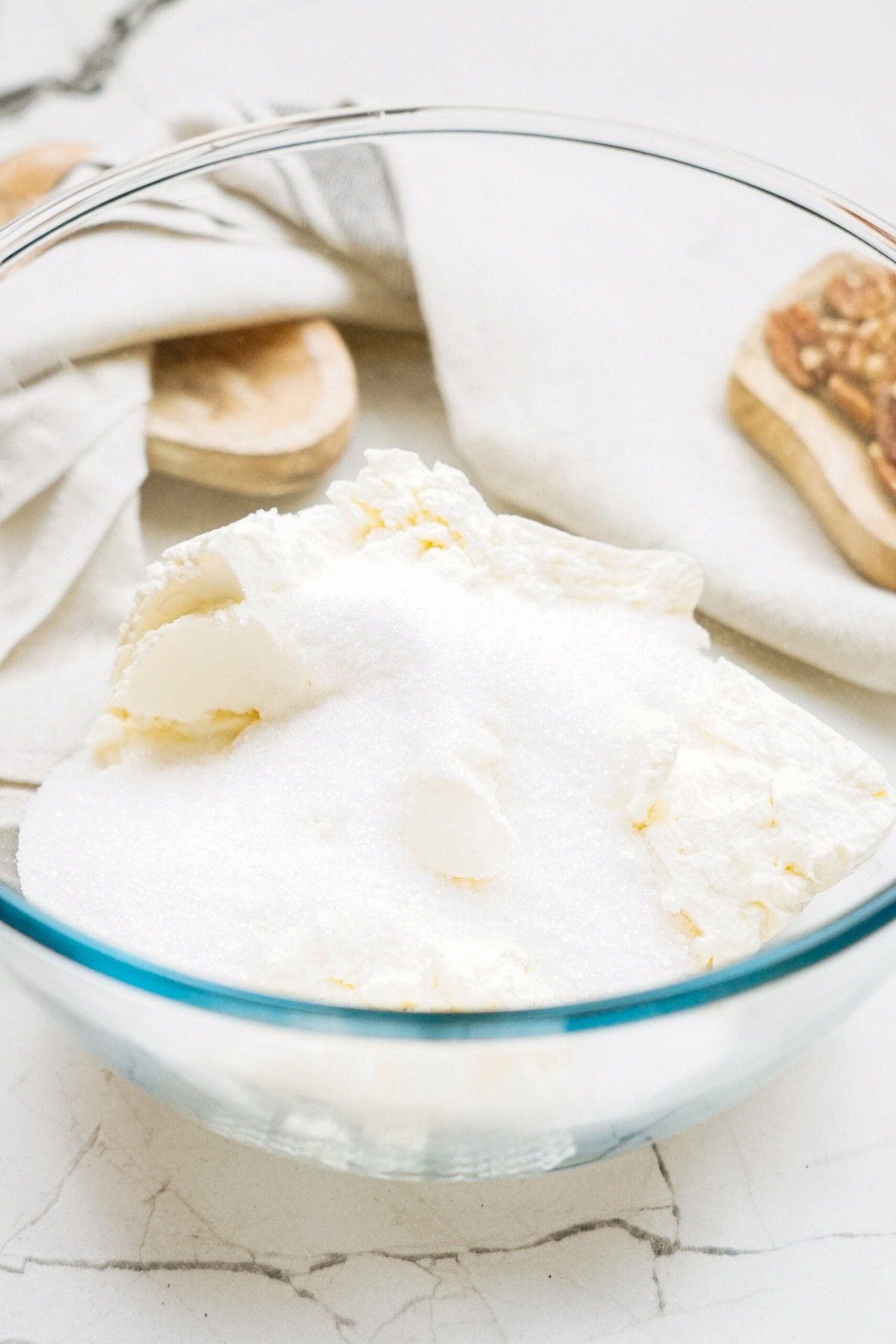 A glass bowl containing sugar and butter on a cracked white surface, with fabric and wooden kitchen utensils in the background.