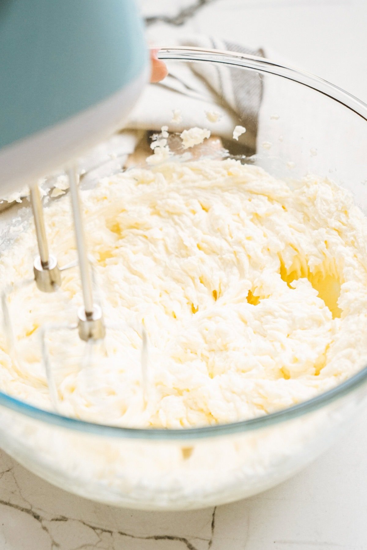 Electric mixer blending creamy batter in a clear glass bowl on a marble countertop.