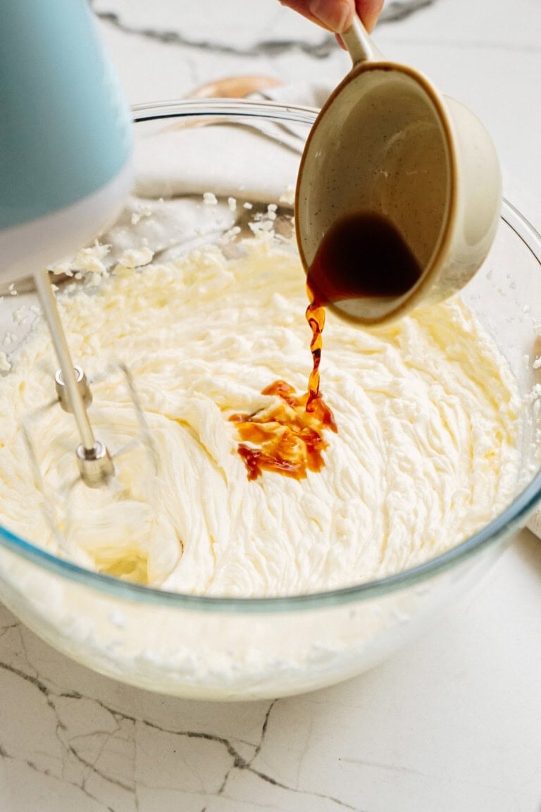 A hand pours vanilla extract into a bowl of whipped cream being mixed by a hand mixer.