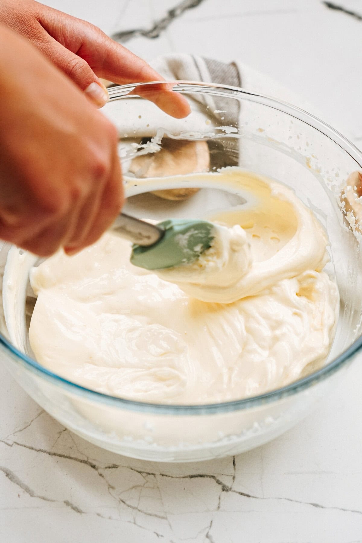 Hands using a spatula to mix creamy batter in a glass bowl on a marble countertop.