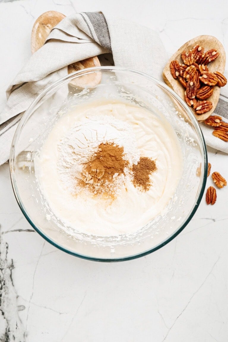 Bowl of batter with flour and spices on top, placed on a marble surface. Pecan nuts and wooden spoons with a cloth are nearby.