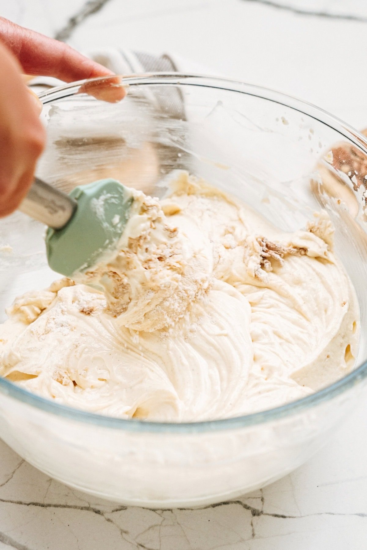 A close-up of a person mixing light-colored batter in a glass bowl with a spatula on a marble countertop.