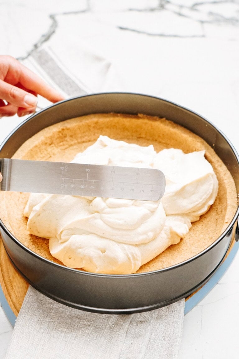 A hand uses a spatula to spread a creamy filling into a pie crust inside a round baking pan.