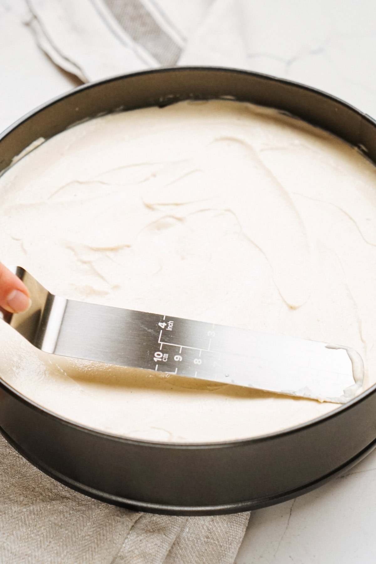 An offset spatula smooths batter in a round baking pan on a light countertop.